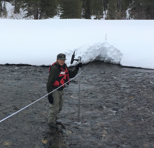 Woman in hip waders, warm clothes and life vest stands in a river taking streamflow measurements.