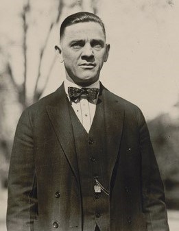 A man in suit stands in the shade of a nearby tree.