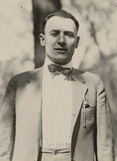 A man in a sit and tie stands in the open air with several trees in the background.