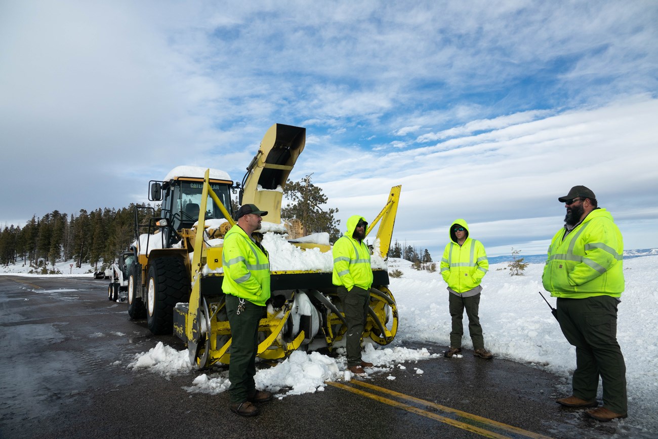 Four men in high visibility jackets stand along a snowy road beside a large yellow snow-removal tractor.