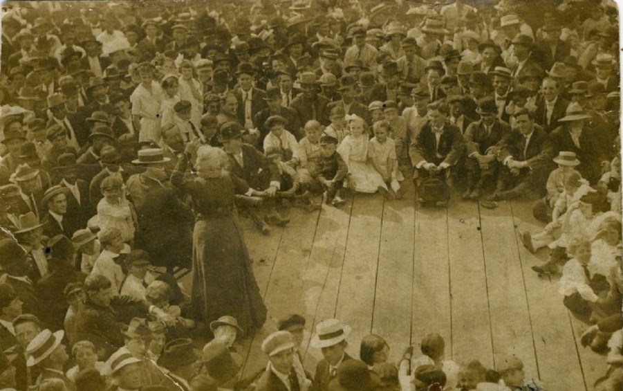 An elderly woman on a wooden stage speaks to a surrounding crowd of men and women seated around her.