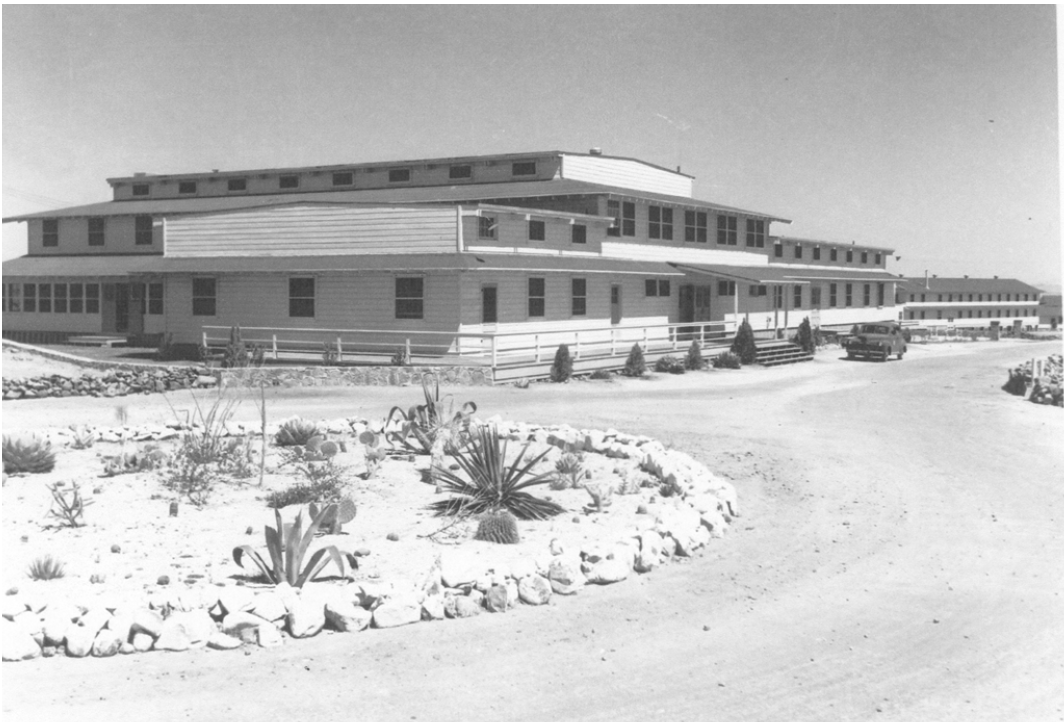 Utilitarian, long and low, two-story wood frame building with several additions that make it appear tiered. It is fronted by a gravel road with some landscaped beds. The beds are surrounded by small boulders and filled with desert plants.