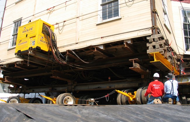 The underside of the corner of the house, lifted up and being moved by large hydraulic lifts on wheels.