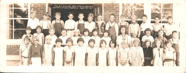 Black and white photo of young students and their teacher. Most of the kids are white, but a handful are of East Asian descent. They stand in three rows in front of a brick wall with windows and open doors.