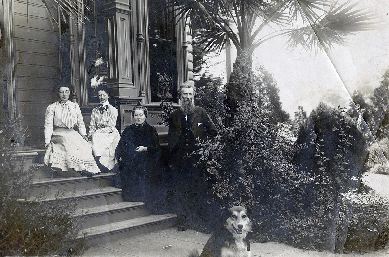 Muir Family Old fashioned portrait of family sitting on porch, two young women in high-necked dresses, older woman in black, and man in suit standing. Dog sits in foreground