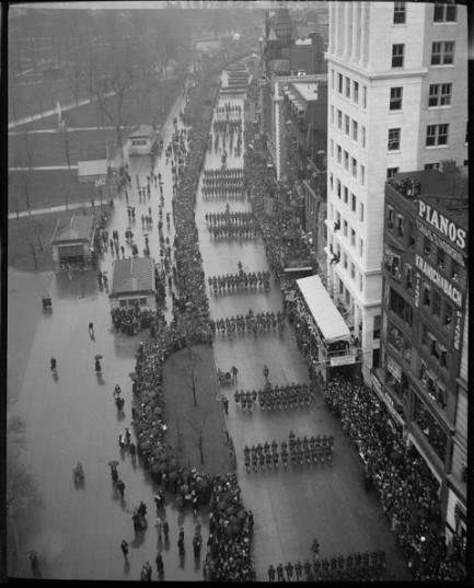 Parade of marching soldiers surrounded by spectators on city streets lined with tall buildings.