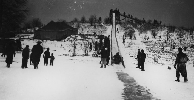 A black-and-white photo looking up a toboggan chute; in the foreground people walk across a snow-covered lake.