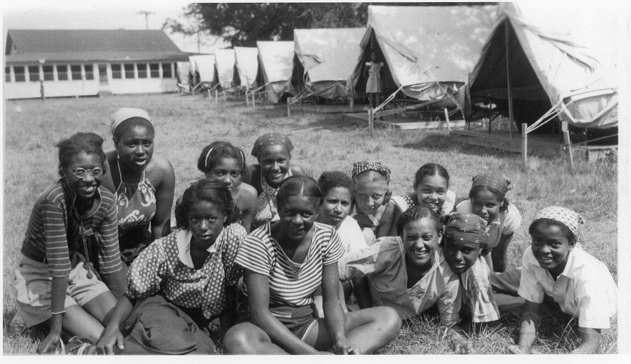 13 African American girls sit on a lawn in front of tents.