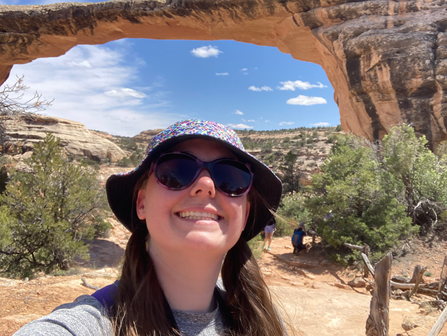A woman wearing a hat and sunglasses stands under a reddish-brown natural rock bridge.
