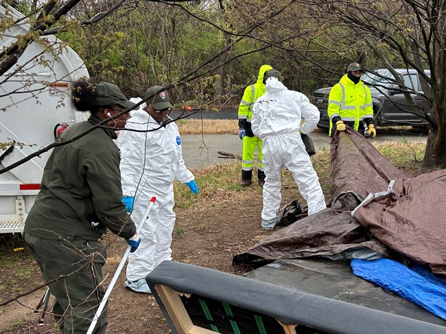 NACE_Encampments Removal_20250324_04_crop Several maintenance workers wearing protective equipment remove tarps and other objects from an unauthorized encampment.