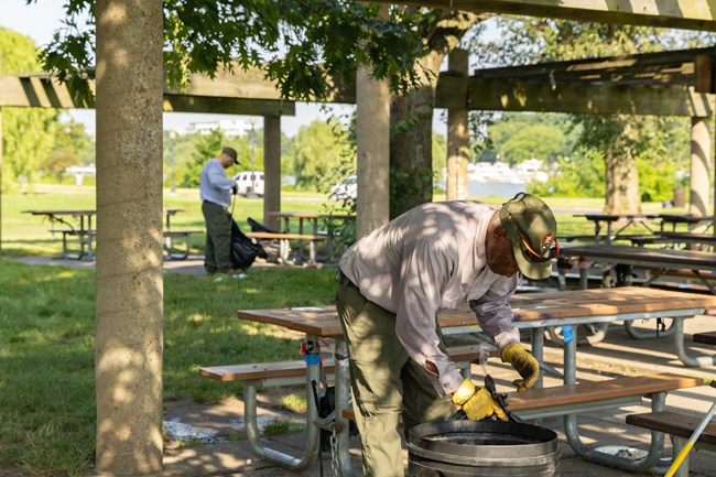 NACE_Trash Pickup_20250622_KGraczyk_130 Two men in NPS uniforms pick up litter among a set of picnic benches in the shade.