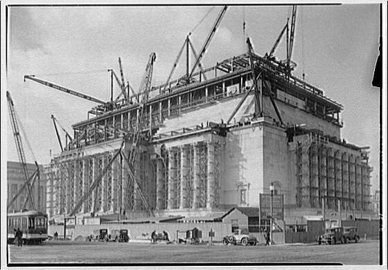 A large building with classical elements being constructed. Large columns line each side of the structure with scaffolding and cranes on the top of the building.
