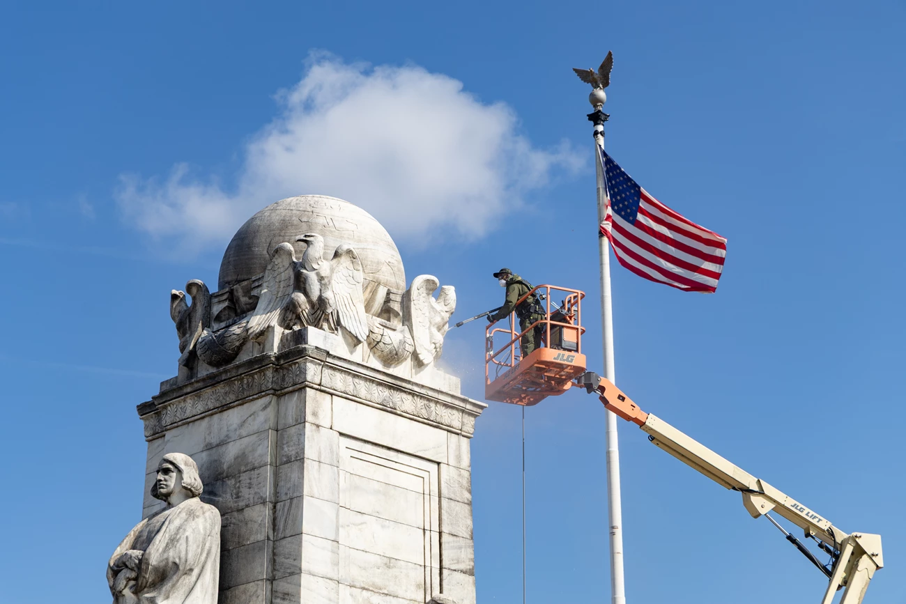 NAMA_Columbus Circle_Clean up_20250708_KGraczyk_104 An NPS maintenance worker stands aloft in a cherry picker and uses a pressure washer to scrub a stone globe with eagles surrounding it, flanked by a large US flag.