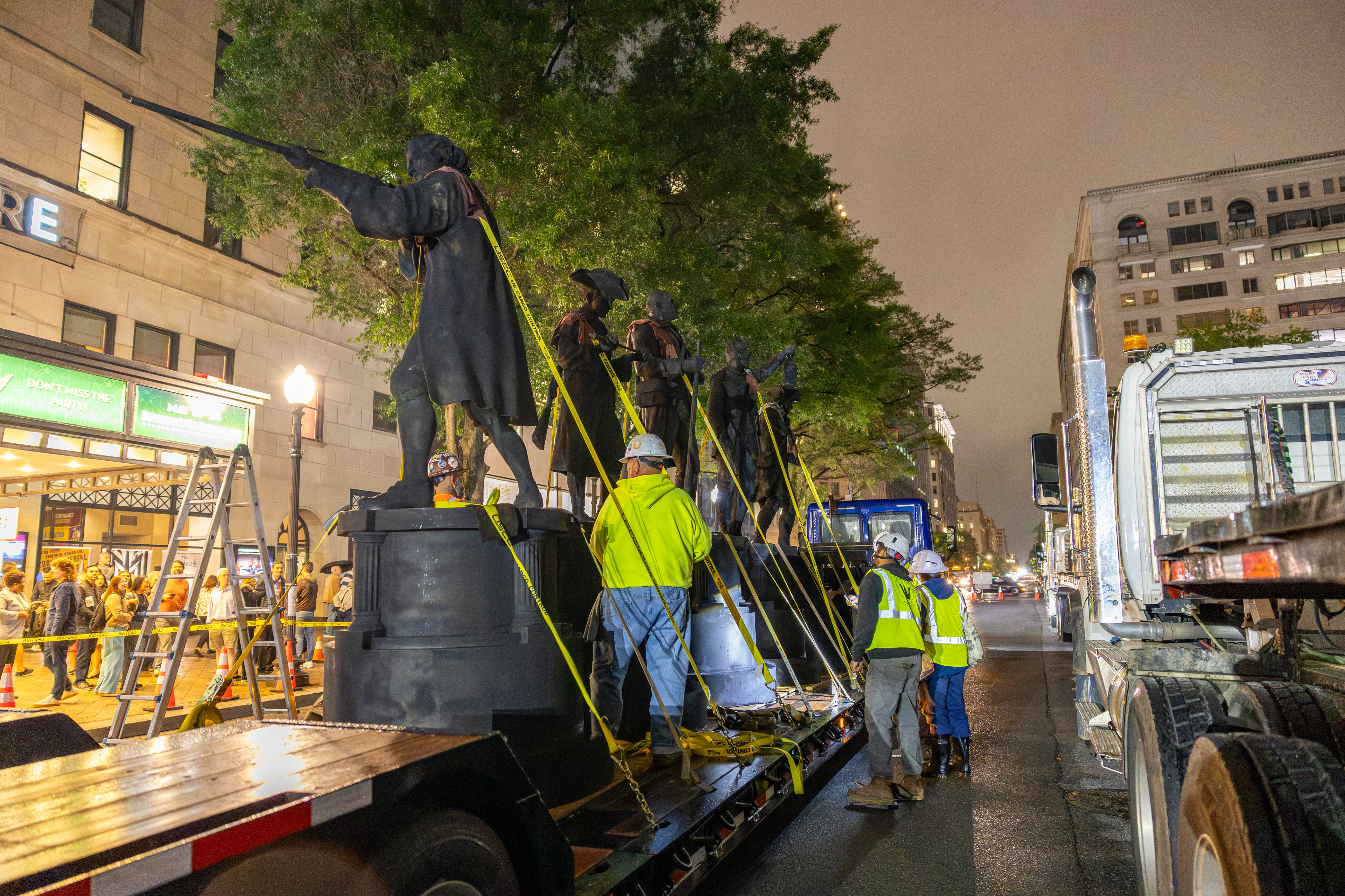 Workers in hard hats and hi-vis vests work to unload several life-sized statues from a flatbed trailer