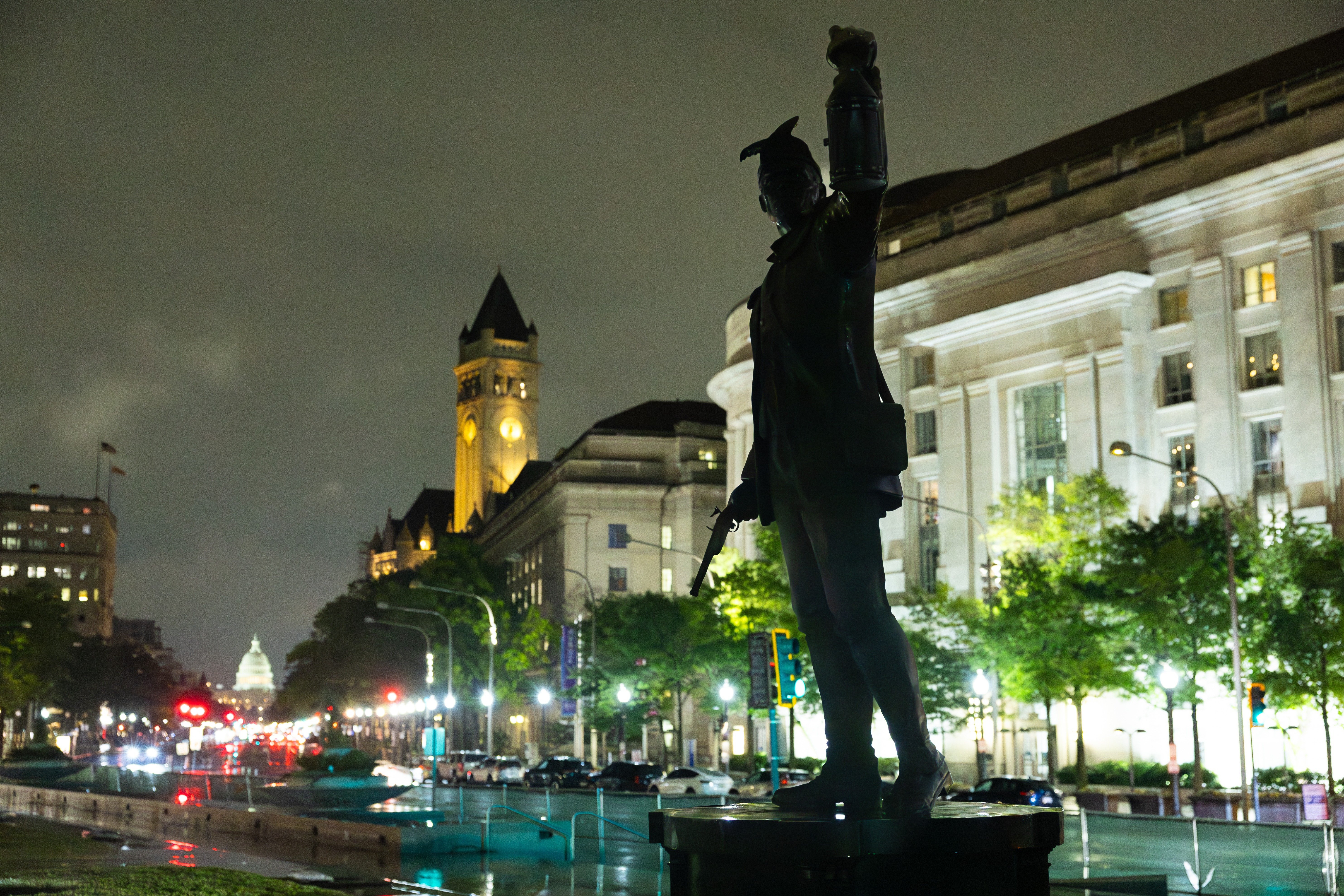 Silhouetted statue of a man holding a pistol and a lantern with a night time city street and the Capitol dome in the background