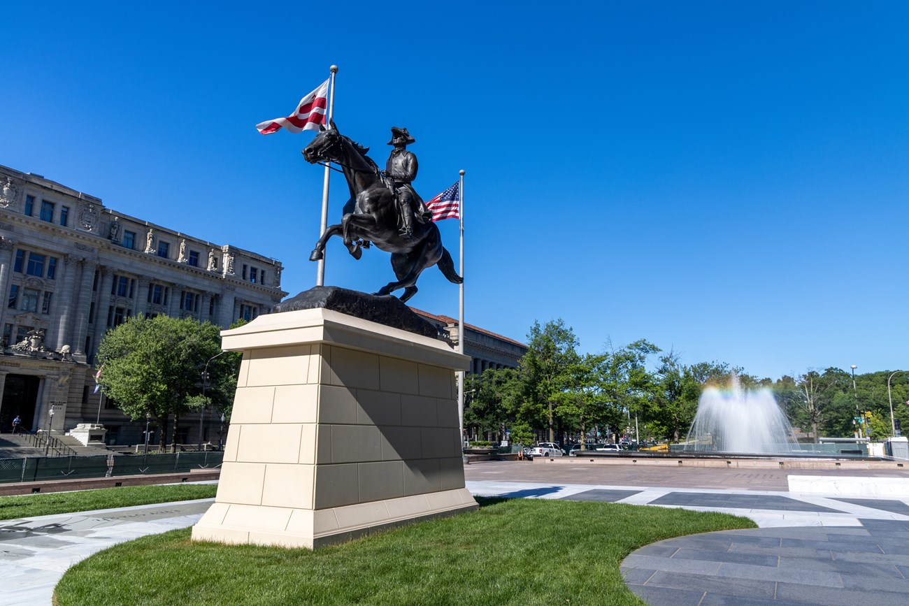 An equestrian statue stands proudly in a square. Behind it, a fountain shoots into the air, creating a rainbow. DC and US flags fly behind the statue.