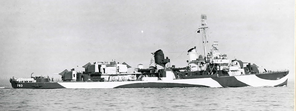 Black and white photograph of a warship in the ocean. Photo views the port broadside of the ship. It is painted with jagged contrasting shapes of dark and light colors as camouflage.