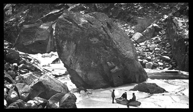 Black and white image of three men sitting on a wooden boat. The boat is on a section of river iced over. Large boulders surround the river.