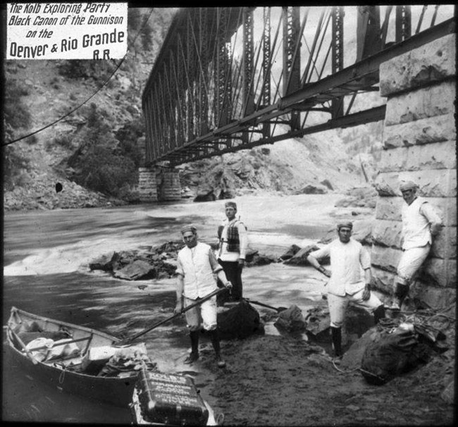 Black and white image of four men standing by a loaded boat and gear bags. They are near the stone base of a truss bridge. Rapids flow under the bridge.