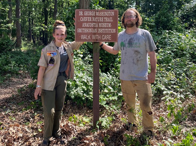 A photo of volunteer Sophie Schell and another volunteer standing next to a park trail sign at National Capital Parks – East.