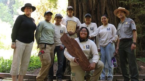 A group of Native American youth interns hold National Park emblems and pose in front of a qiant sequoia.