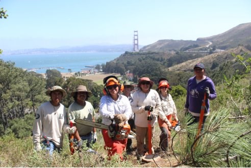 A group of Native American youth stand on a brushy hillside with the San Francisco Bay Bridge in the distance.