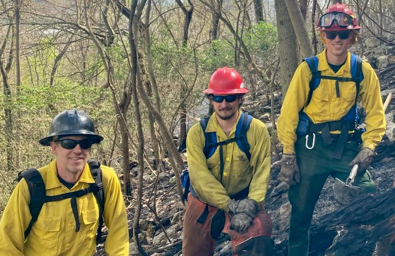 Wildland firefighters from across the region come together from different parks to help suppress fires. Three firefighters stand in a recently burned area and smile at the camera.