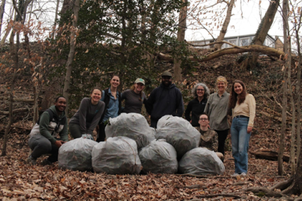 A group of volunteers pose for a photo in the woods, they stand behind a pile of trash bags filled with debris, fallen leave cover the ground.