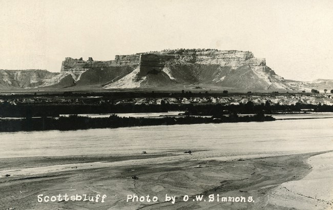 A black and white photo shows a wide river flowing past a large, sandstone bluff.