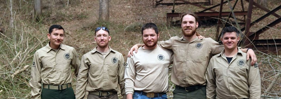 New River Gorge National Park and Preserve Veterans Fire Crew Five smiling men in beige uniform shirts stand at base wooded slope.