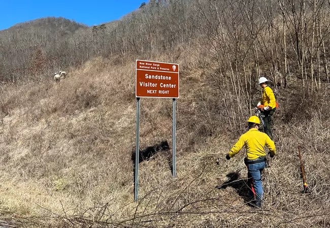 Two people in wildland fire gear stand near a sign which reads Sandstone Visitor Center next right.