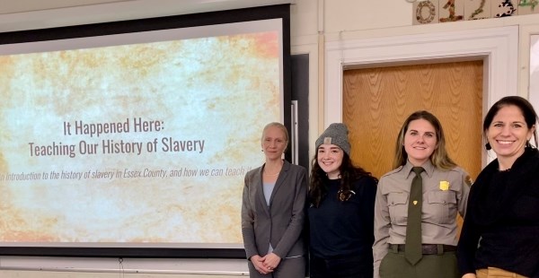 A photo of four women next to a projected screen with a slide that reads "It Happened Here: Teaching Our History of Slavery"