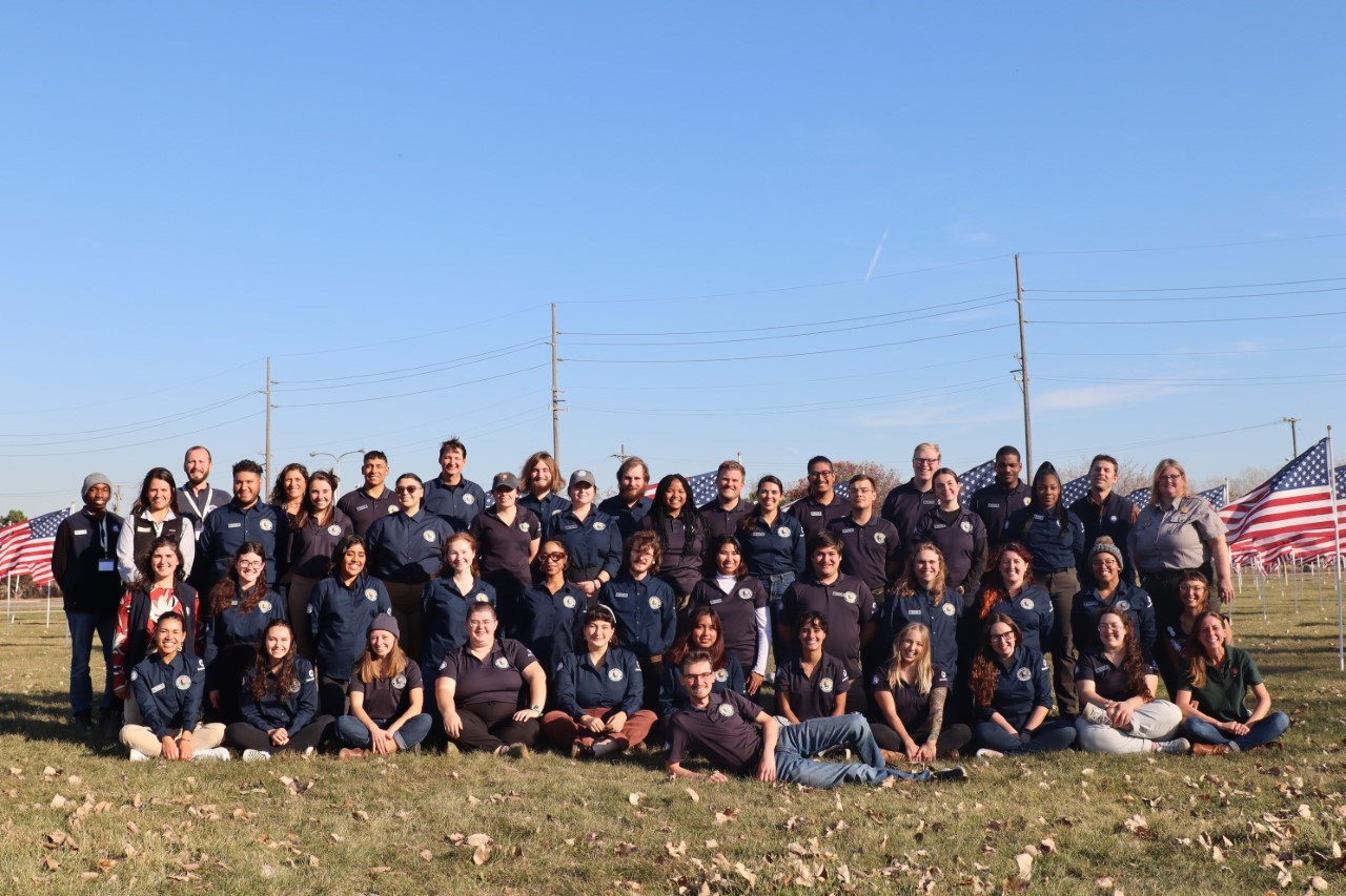 Group photo of Community Volunteer Ambassadors, National Park staff, and Conservation Legacy staff outdoors in a field of United States of America flags