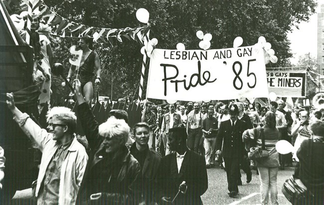 Archive photo of a Lesbian and Gay Pride March in 1985. Shows many people marching and holding signs that say, "Lesbain and Gay Pride '85"