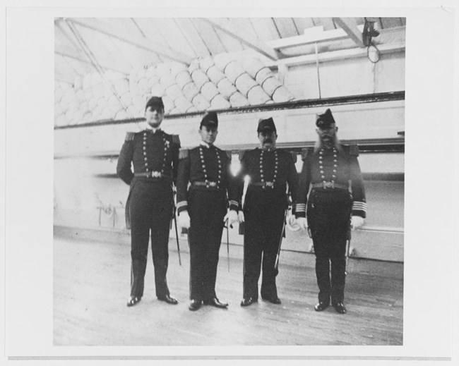 Four officers in early 1900s naval uniforms standing on the deck of a ship.