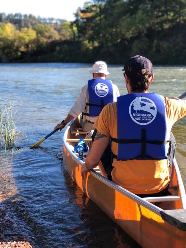 Two people wearing blue life jackets that say "Niobrara Volunteer" launch a canoe into the river