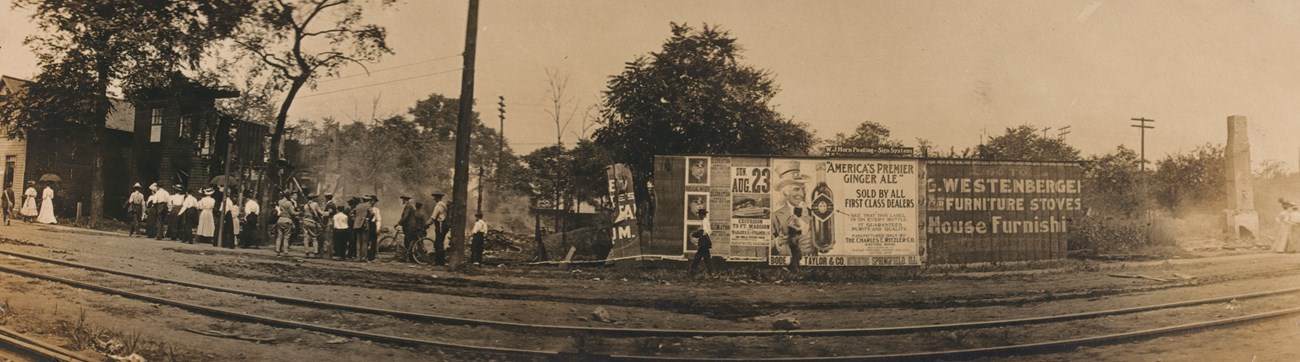 A row of smoldering and charred house frames line a city street. Onlookers and soldiers stand on the sidewalk, looking at the houses.