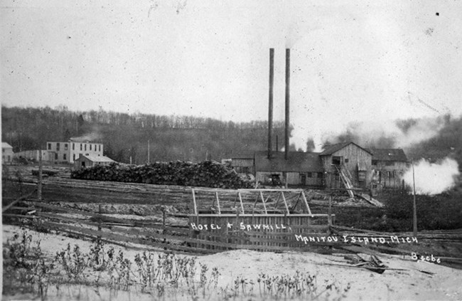 NMI Crescent Mill A black and white photo of a sawmill situated in a wide, sandy area before a large tree covered hill. There is smoke billowing out of the mill. The ground is covered in iron tracks, and beside the mill are vast piles of milled lumber.