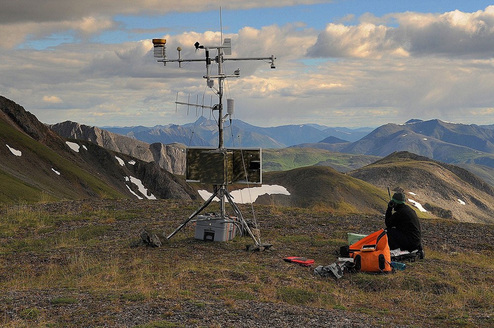 Climate Station a person sits next to equipment for monitoring climate on the top of a mountain with mountains in the distance