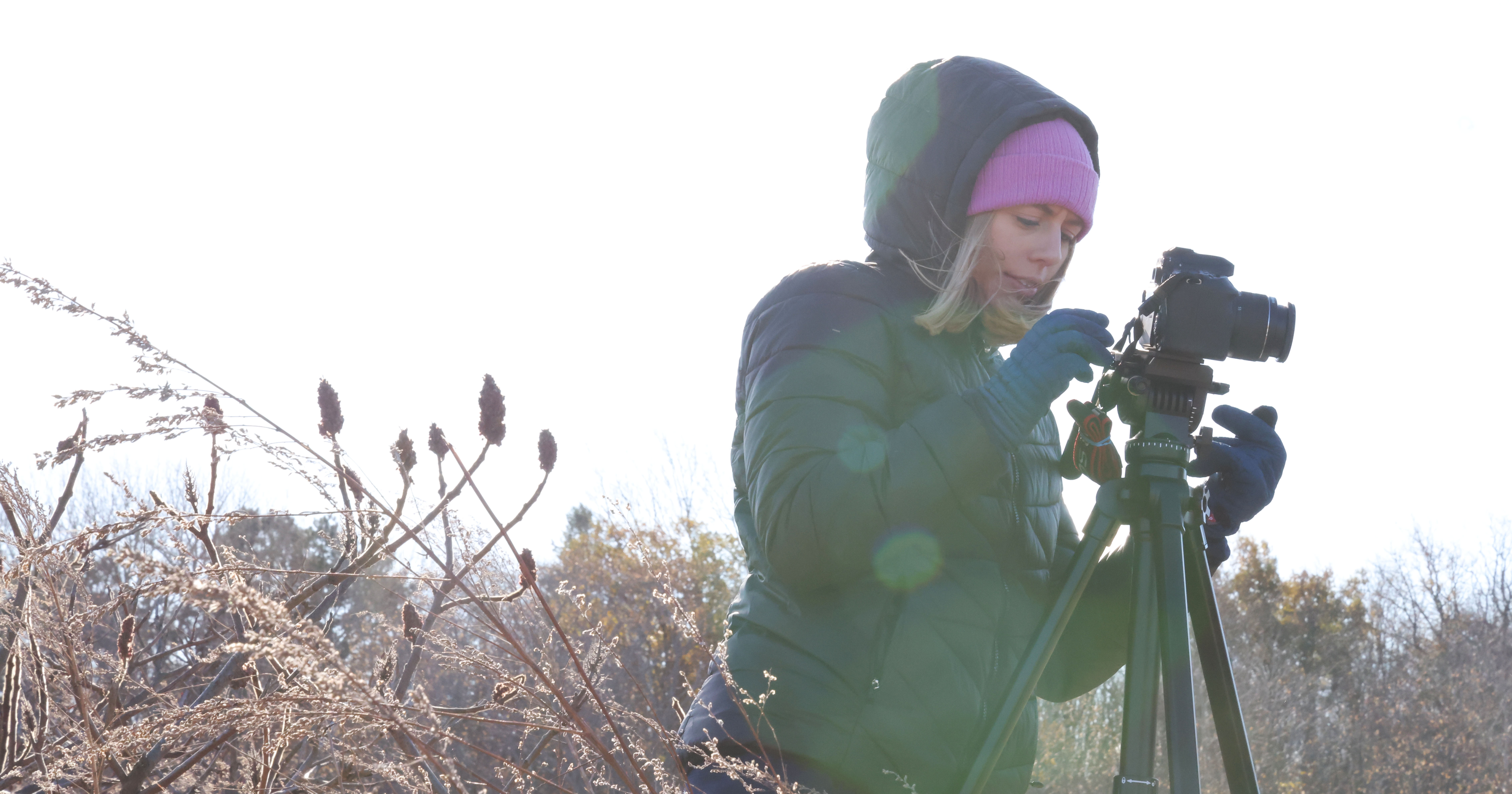 A person in a black coat, gloves, and a purple hat adjusts the settings on a camera attached to a tripod. The sky is grey and there are reeds and low shrubs in the foreground.