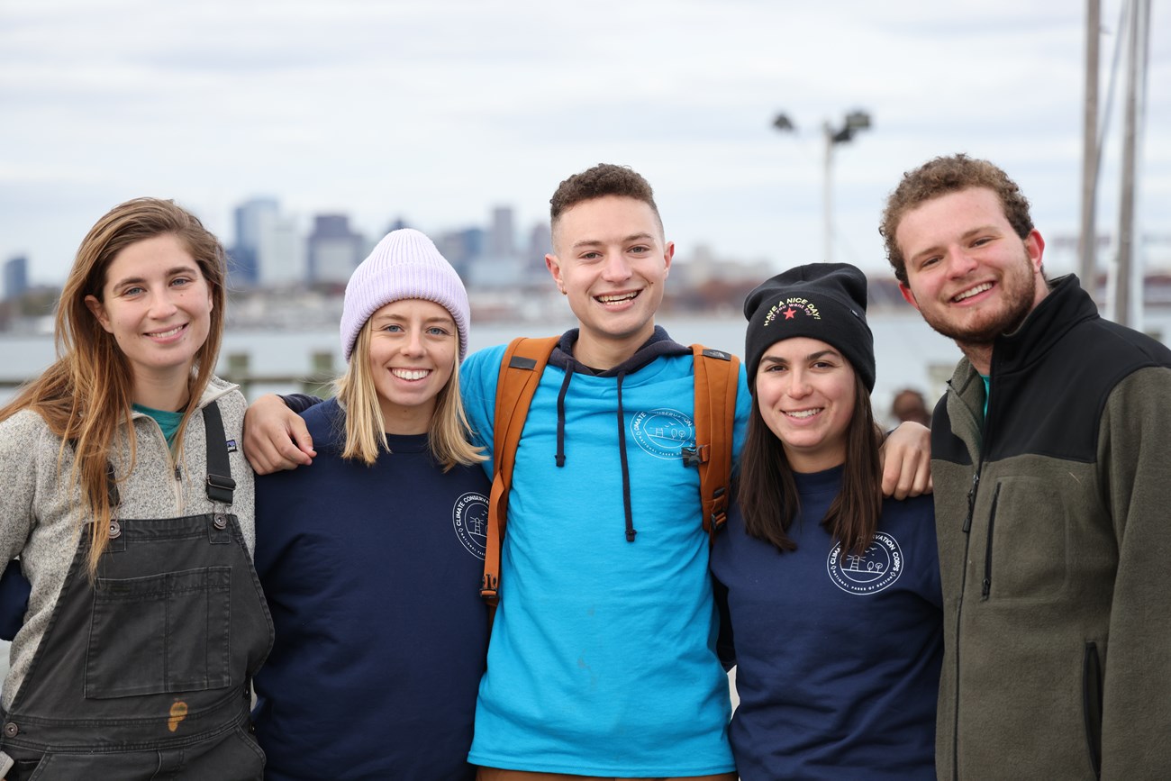 A group of five young adults with their arms around each other smiling at the camera. They are outside on an island overlooking Boston Harbor, with the city skyline in the background.