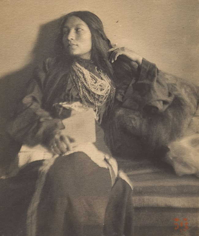Seated woman with long hair and bead necklaces looks to her right