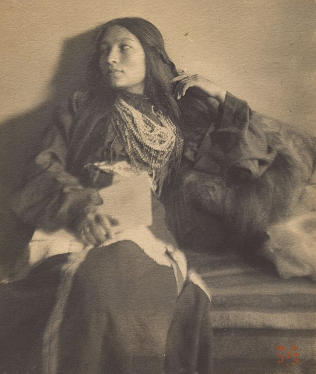 Zitkála-Šá, 1898. Photo by Joseph Turner Keiley. Seated woman with long hair and bead necklaces looks to her right