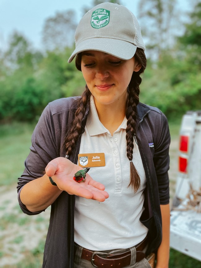 A woman wearing a tan shirt and hat with the Fish & Feathers logo holds a small green and red bird.