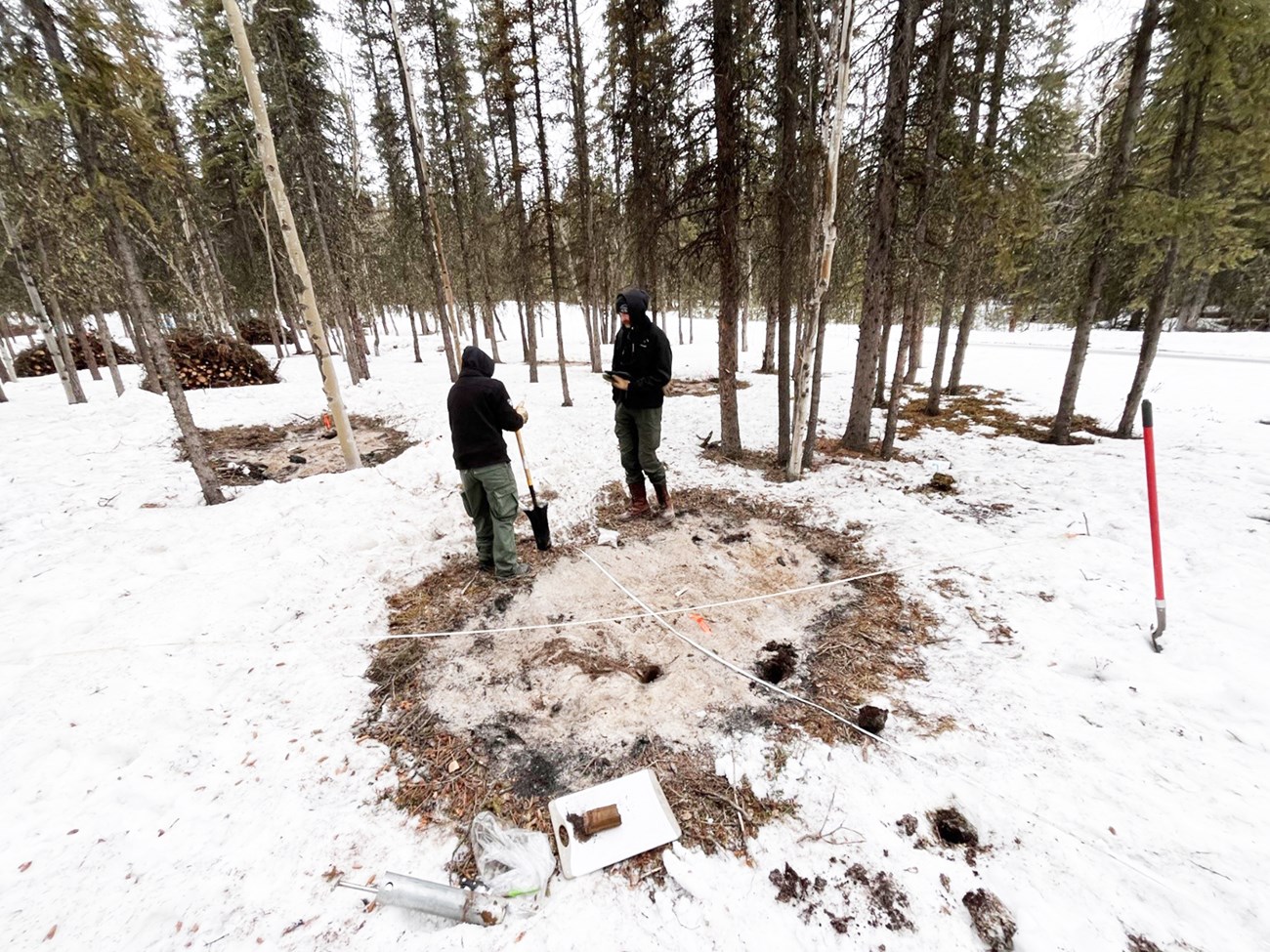 Two people stand near a burned area on snow, with tape laid down in a cross pattern over the area