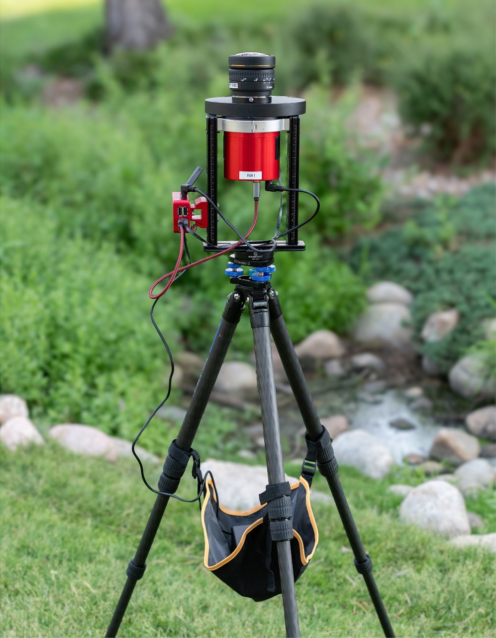 Camera on tripod with wires attached to it standing on green grass in front of a rocky stream