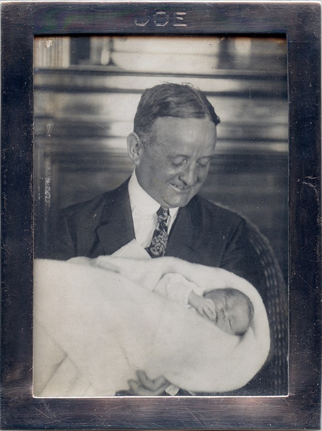 A framed black and white photo of John F. Fitzgerald holding Joseph P. Kennedy Jr.  Fitzgerald is wearing a dark suit.  At the top of the frame, the name "Joe" is engraved.