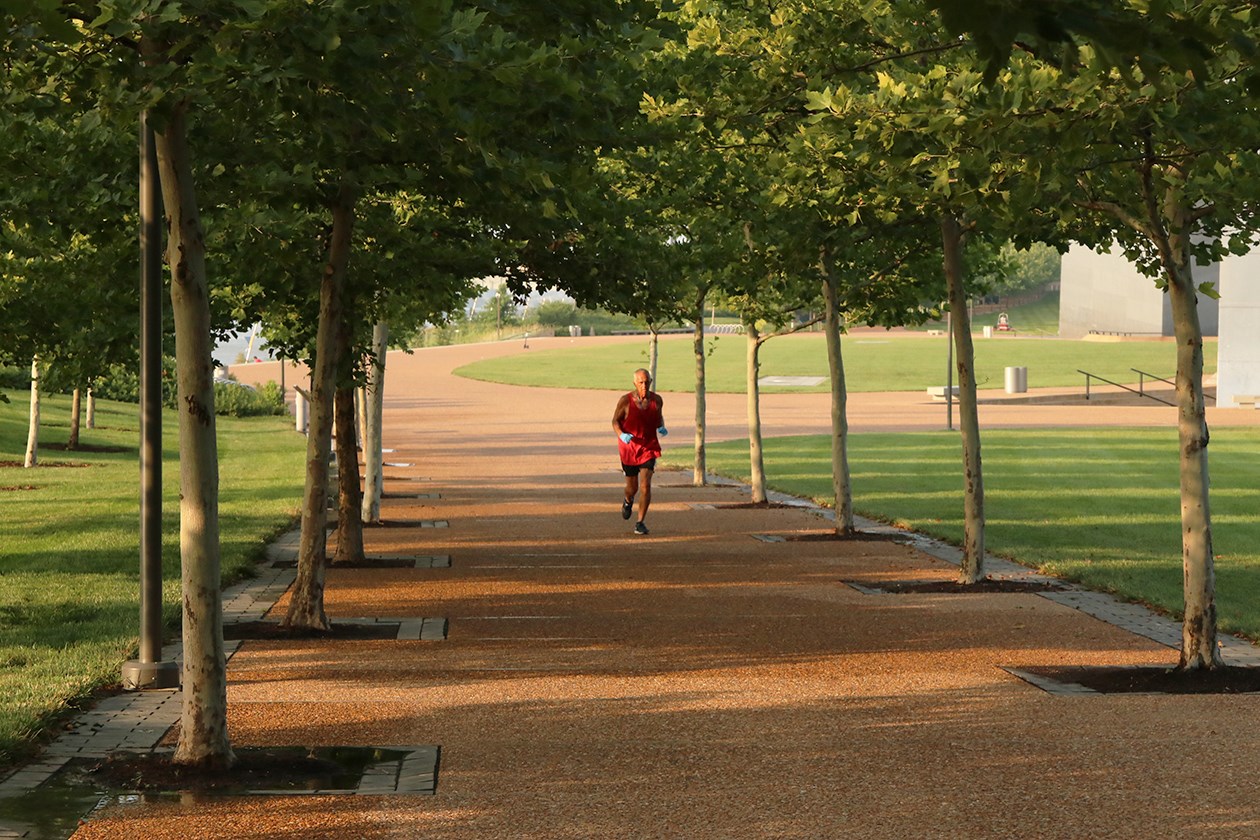 A man wearing a red tank top and black shorts runs on a path lined with landscaping trees at Gateway Arch National Park