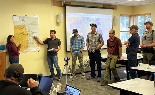Several people gather around a map taped to a wall which two people are pointing to and discussing