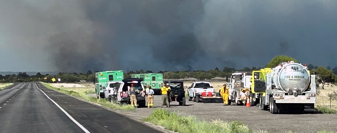 The Encerrita Fire pushes into an area partially treated by prescribed fire. Firefighters in a pullout with fire vehicles while dark smoke rises in the near distance in pinyon-juniper forest.